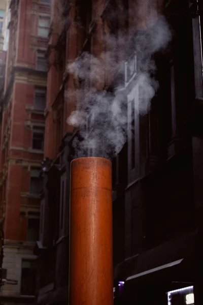 Tubo de ventilación humeante en la calle cerca de edificios de piedra sobre fondo borroso en la ciudad de Nueva York - foto de stock