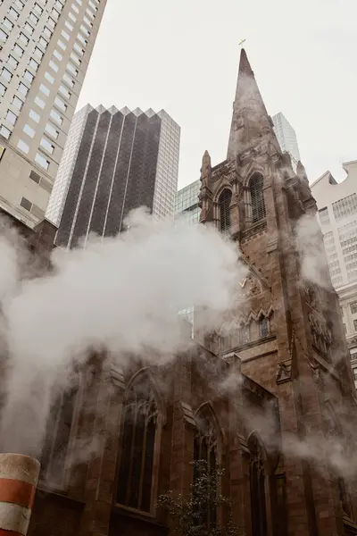 Vista de ángulo bajo de la iglesia católica de ladrillo rojo cerca de rascacielos y vapor en la calle de la ciudad de Nueva York - foto de stock