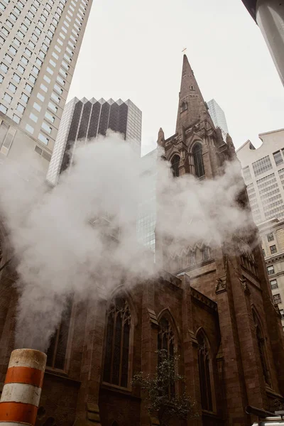 Iglesia católica de ladrillo rojo y rascacielos cerca de vapor en la calle en la ciudad de Nueva York, vista de ángulo bajo - foto de stock