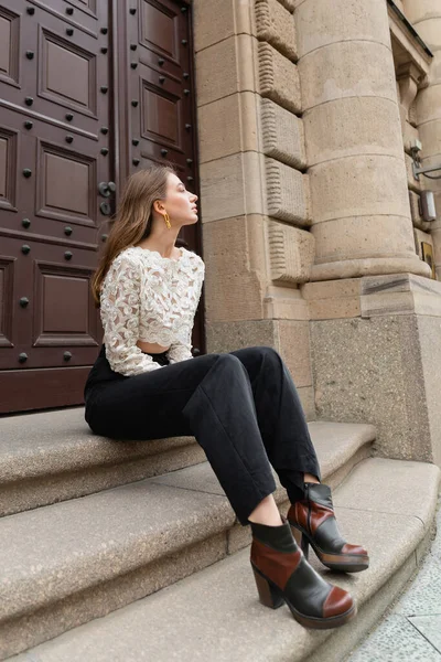 Модна молода жінка в білому мереживі зверху і високі талії штани, що сидять на сходах біля дверей будівлі — стокове фото