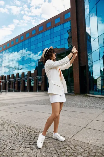 Eine Frau im schicken weißen Outfit macht ein Selfie vor einem modernen Gebäude in einer europäischen Stadt. — Stockfoto