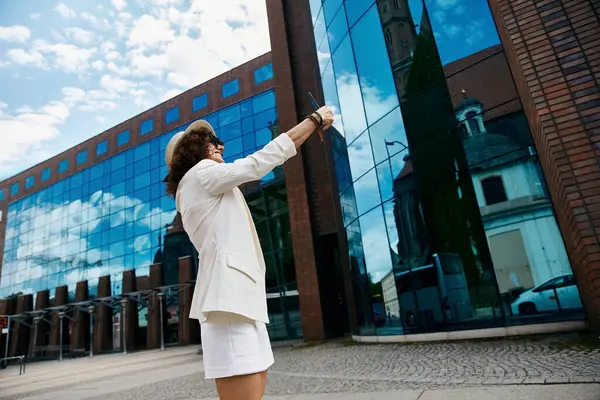 A woman in a white blazer and skirt takes a selfie in front of a modern building with a reflection of a European city. — Stock Photo