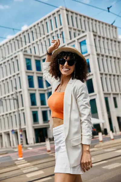 A stylish woman strolls down a city street in Europe, wearing a white blazer and orange top. — Stock Photo