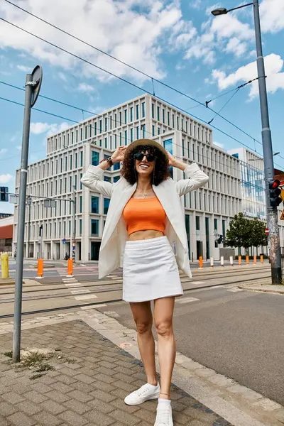 Una donna in un blazer bianco e top arancione cammina con fiducia su una strada della città. — Foto stock