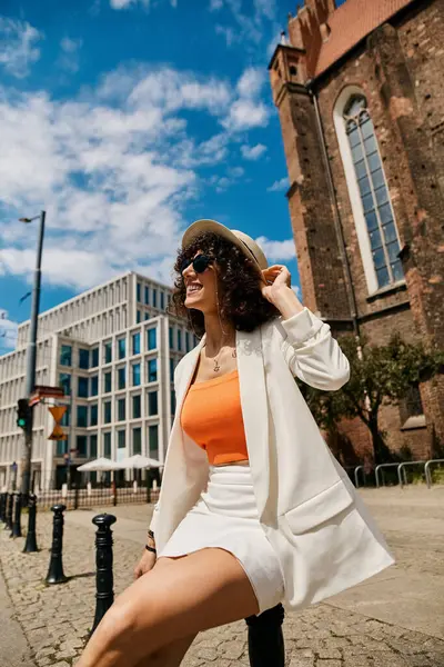 Eine stilvolle Frau in weißem Blazer und orangefarbenem Oberteil genießt einen sonnigen Tag in Europa. — Stockfoto