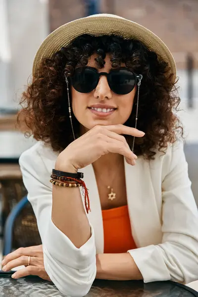 A woman in a white blazer and sunglasses sits at a cafe, enjoying the sights and sounds of Europe. — Stock Photo