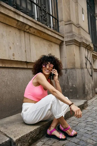 A woman in pink and white outfit sits on a step in a European city, talking on her phone. — Stock Photo