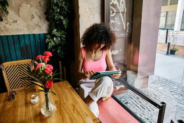 Eine Frau im rosafarbenen Tanktop sitzt am Fenster und benutzt ein Tablet. — Stockfoto