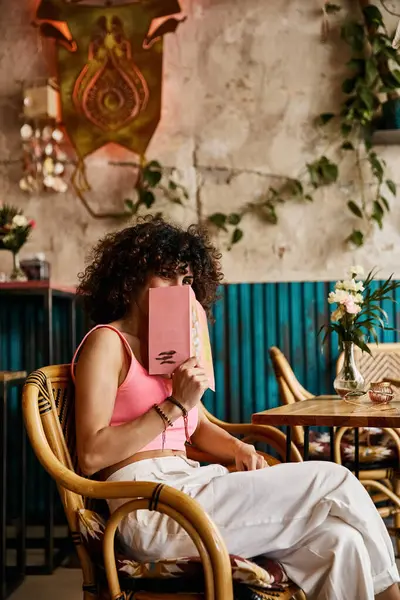 A woman in a pink top and white pants sits in a cafe, reading a book while enjoying the European ambiance. — Stock Photo