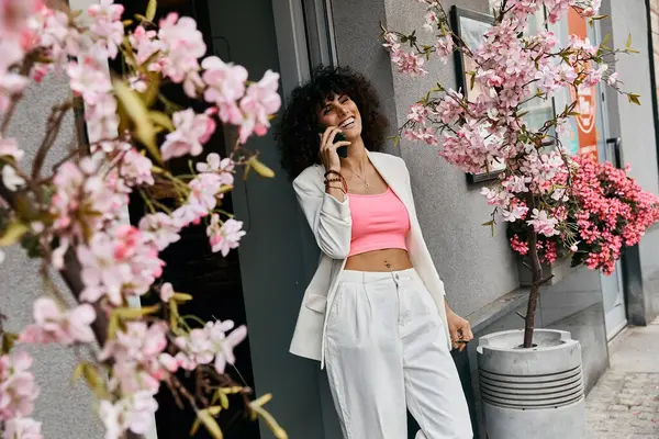 Eine stylische Frau in weißem Blazer und rosa Top lächelt auf ihrem Handy in einer europäischen Stadt. — Stockfoto