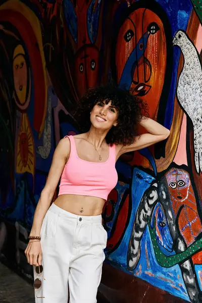 A stylish woman in a pink top and white pants poses against a colorful mural in Europe. — Stock Photo