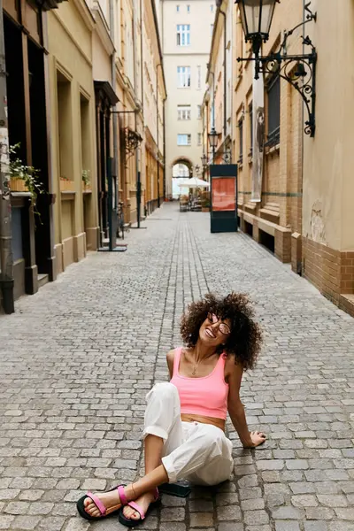 Eine stilvolle Frau genießt einen sonnigen Tag in einer europäischen Stadt und lacht, während sie auf einer Kopfsteinpflasterstraße sitzt. — Stockfoto
