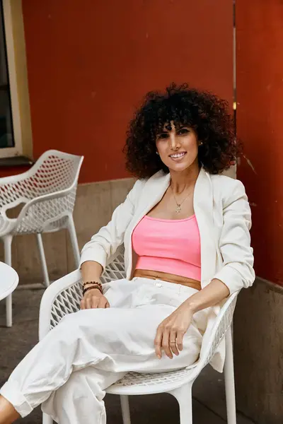 Eine stilvolle Frau in weißem Blazer und rosa Oberteil sitzt auf einem Stuhl in einer europäischen Stadt und genießt einen entspannten Moment. — Stockfoto