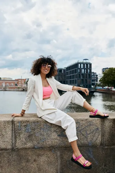 Eine Frau in weißem Blazer und weißer Hose posiert auf einer Steinmauer mit Blick auf einen europäischen Kanal. — Stockfoto