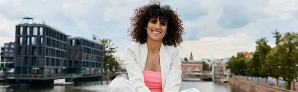 Uma mulher em um blazer branco sorri brilhantemente enquanto se senta em uma ponte com vista para um canal europeu. — Fotografia de Stock