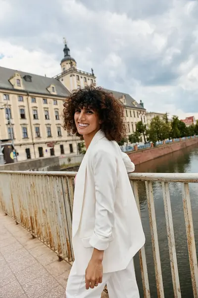 A woman in a white blazer smiles while walking on a bridge in a European city. — Stock Photo