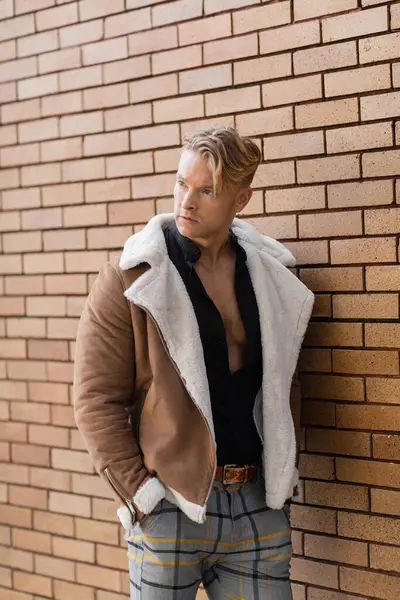 A handsome blonde man in a stylish brown jacket and plaid pants stands against a brick wall — Stock Photo