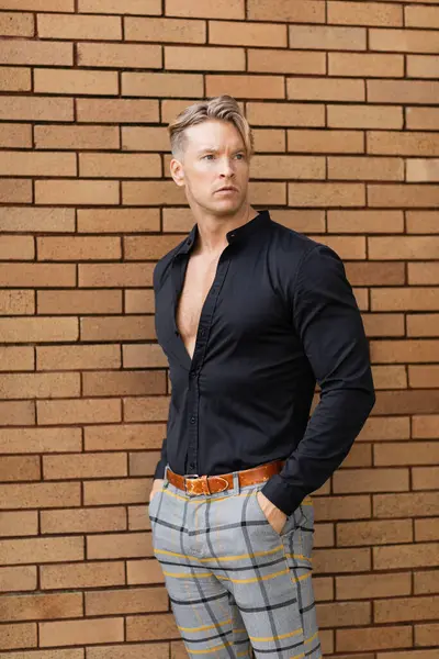 A handsome blonde man stands on a brick street. He is wearing a stylish black shirt with the top button undone and plaid pants. — Stock Photo