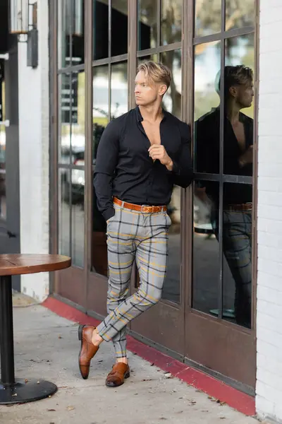 A handsome blonde man wearing a black shirt, plaid pants, and brown shoes leans against a doorway in Orlando, Florida. — Stock Photo