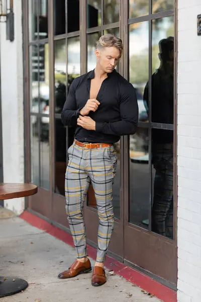 Un guapo hombre rubio con camisa negra y pantalones a cuadros se encuentra frente a un edificio en Orlando, Florida. - foto de stock