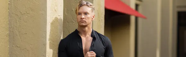 A handsome blonde man in stylish attire stands on a street in Orlando, Florida. He is leaning against a building with his shirt unbuttoned. — Stock Photo