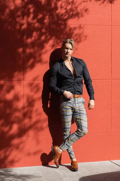 Un guapo hombre rubio con un atuendo elegante se apoya contra una pared roja en Orlando, Florida, bañado en el cálido sol de la tarde. - foto de stock