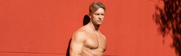 Un uomo muscoloso, senza maglietta, con i capelli biondi posa davanti a un muro rosso. — Foto stock