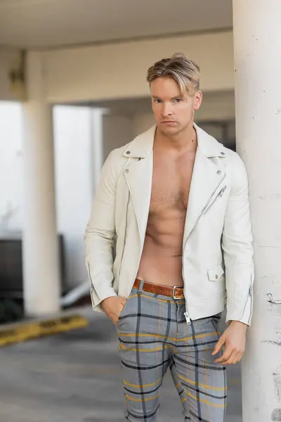 A handsome blonde man in a white leather jacket and plaid pants poses against a pillar in Florida. — Stock Photo