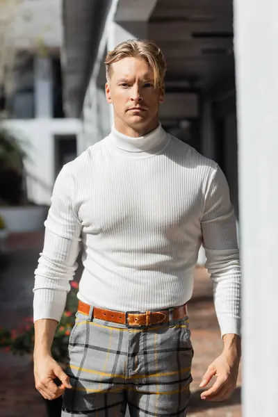 Un bel homme blond pose dans un pull à col roulé blanc et un pantalon à carreaux sur une rue ensoleillée de Floride. — Photo de stock