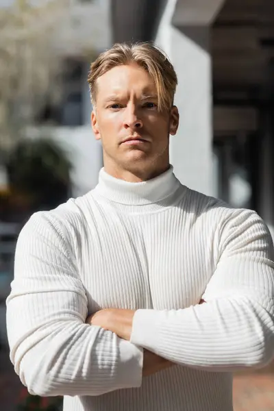 Un guapo hombre rubio posa en una calle de Florida, vistiendo un jersey de cuello alto blanco y mirando directamente a la cámara. - foto de stock