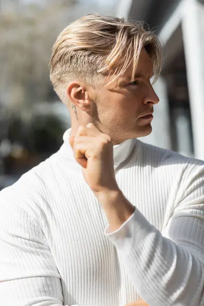 Un guapo hombre rubio con un corte de pelo elegante posa en una calle de Florida, usando un suéter de cuello alto blanco. — Stock Photo