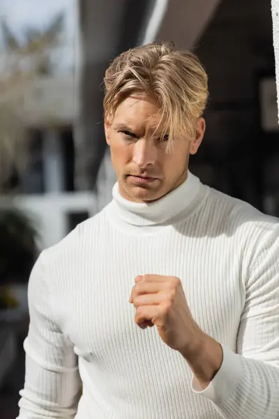 A handsome blonde man with a determined expression poses for a photo while wearing a white turtleneck sweater on a Florida street. — Stock Photo