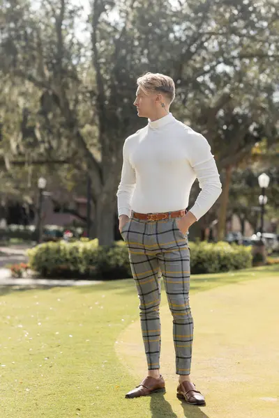 Un guapo hombre rubio con un atuendo elegante camina con confianza a través de un exuberante parque verde en Miami, disfrutando del sol. - foto de stock