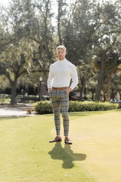 A blonde man wearing a white turtleneck and checkered pants stands on a green lawn in Miami. — Stock Photo
