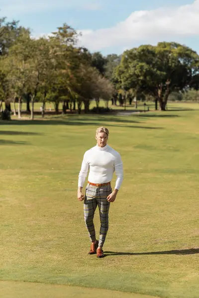 Un uomo biondo con un dolcevita bianco e pantaloni a quadri cammina in un parco verde a Miami. — Foto stock