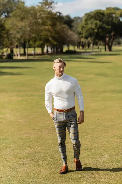 Un uomo biondo con un dolcevita bianco e pantaloni a quadri attraversa un parco erboso a Miami. — Foto stock