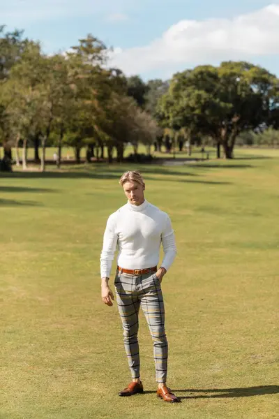 A blonde man in a white turtleneck and patterned pants walks through a lush green park in Miami. — Stock Photo