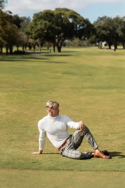 Un uomo biondo con un dolcevita bianco e pantaloni scozzesi siede sull'erba verde di un campo da golf di Miami. — Foto stock