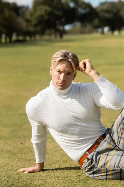 Un uomo biondo elegante in un dolcevita bianco e pantaloni a quadri posa su un parco erboso a Miami. — Foto stock