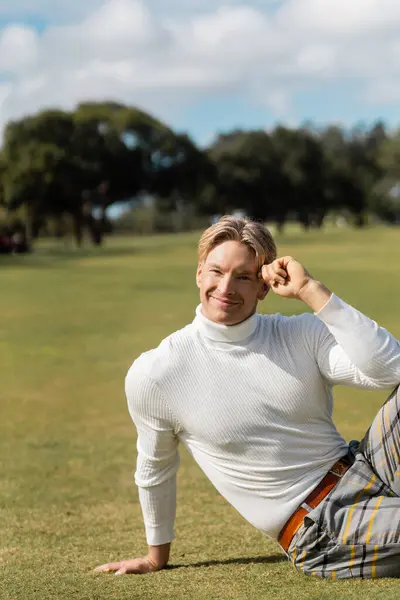 A good-looking blonde man in stylish attire smiles while sitting on the green grass of a Miami park. — Stock Photo