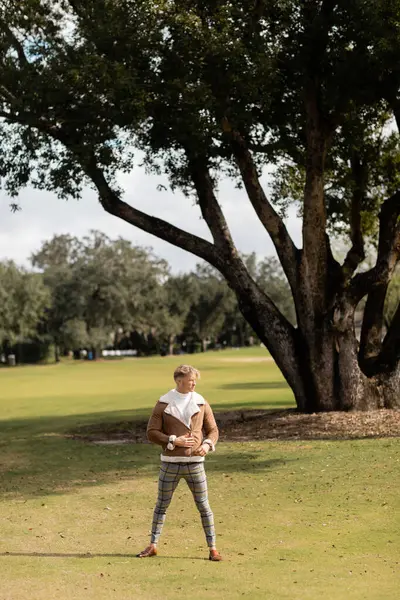 Un uomo biondo in giacca marrone e pantaloni a quadri cammina attraverso un parco verde a Miami. — Foto stock