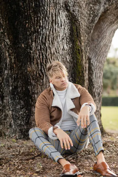 A blonde man in stylish attire leans against a tree in a green Miami park. — Stock Photo