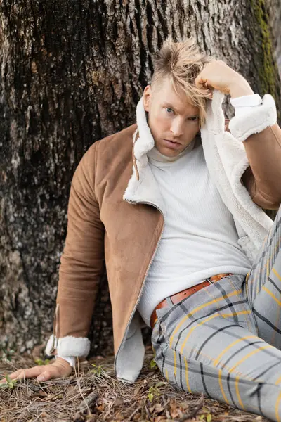 A handsome blonde man wearing a brown suede jacket and plaid pants in park near tree — Stock Photo