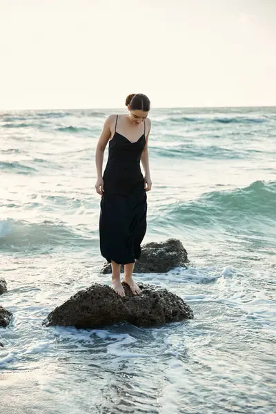 A young woman in a black sundress stands on a rock in the surf as the sun sets over Miami Beach. — Stock Photo