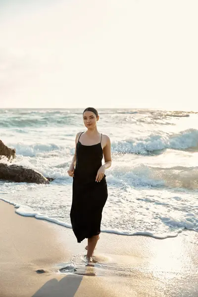 A girl in a black sundress walks Miami sandy beach, ocean waves cresting behind her. — Stock Photo