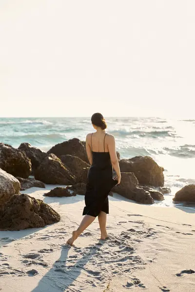 A young woman in a black sundress walks along a sandy Miami beach, gazing out at the ocean. — Stock Photo