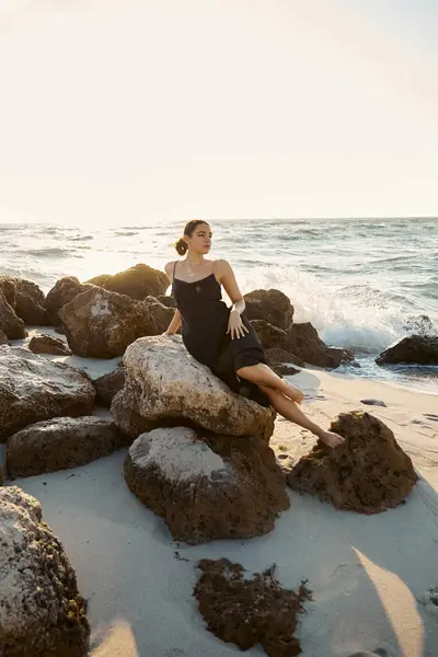 A young woman in a black sundress sits on a rock by the ocean, enjoying the warm Miami sunshine. — Stock Photo