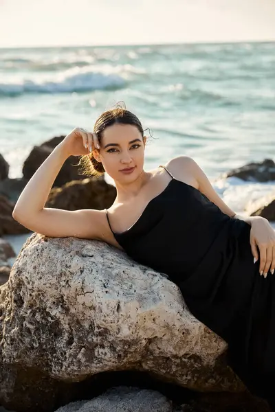 A young woman in a black sundress leans against a rock on a Miami beach, gazing at the ocean. — Stock Photo