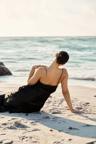 A woman in a black sundress sits on the sand, gazing out at the ocean. The setting sun casts a warm glow on the scene. — Stock Photo