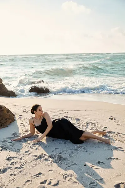Uma jovem mulher de vestido preto jaz na areia branca de Miami Beach, olhando para o oceano azul-turquesa.. — Fotografia de Stock